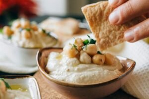 Mão mergulhando uma torrada em uma pasta de homus com grão de bico em cima